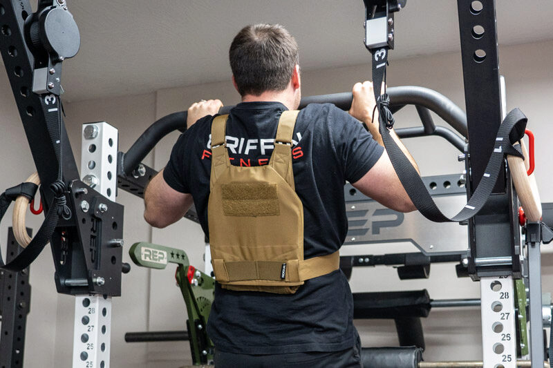A man utilizes a pull-up bar in a gym setting, evaluating the performance of the Rogue Plate Carrier during his workout.