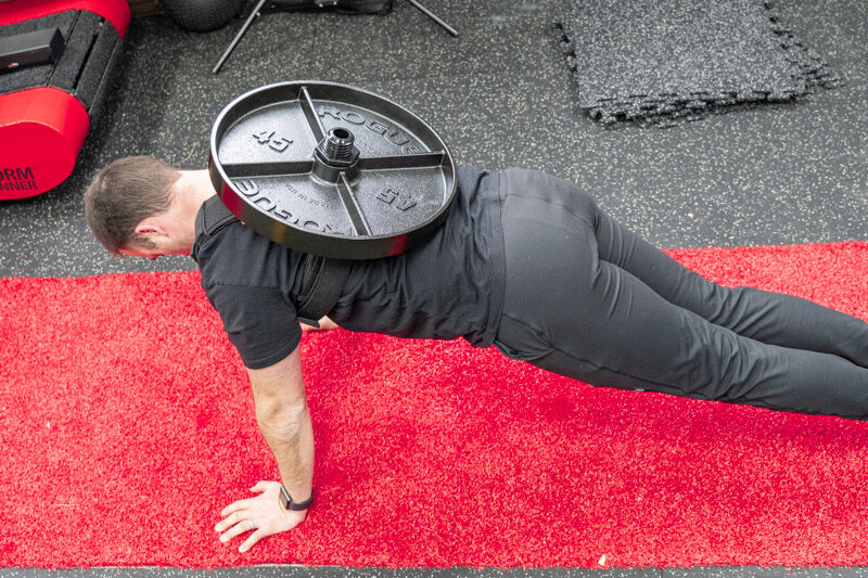overhead shot of a man holding a plank position on a red and black gym mat, wearing the Kensui EZ-VEST Plate-Loadable Weight Vest with two 45 lb. Rogue Fitness weight plates attached.
