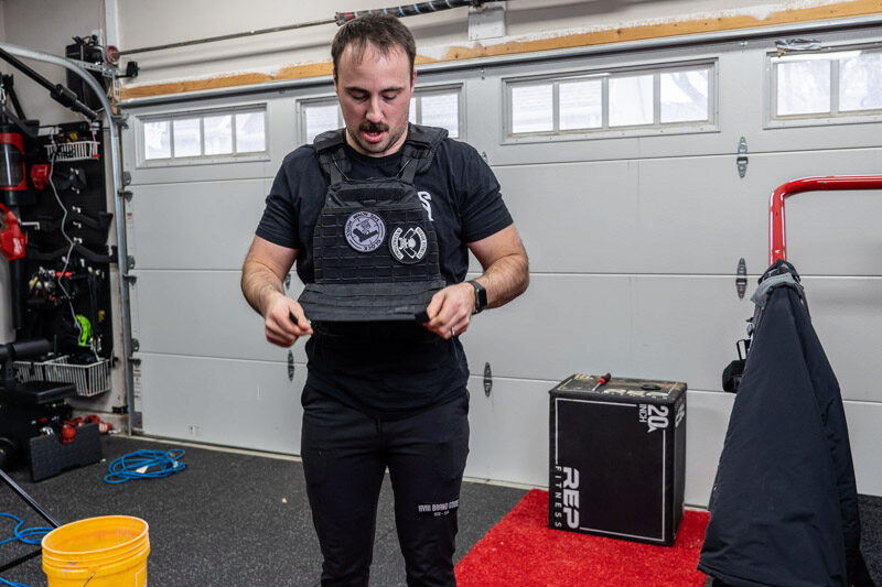 A man wearing the black 5.11 TacTec Plate Carrier in a home garage gym, while holding the edge of the vest and analyzing its quality.