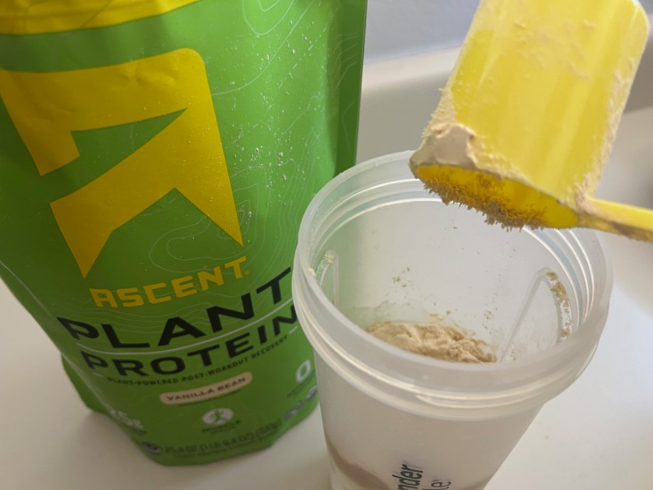 A scoop of Ascent Plant Protein is being poured into a shaker cup.