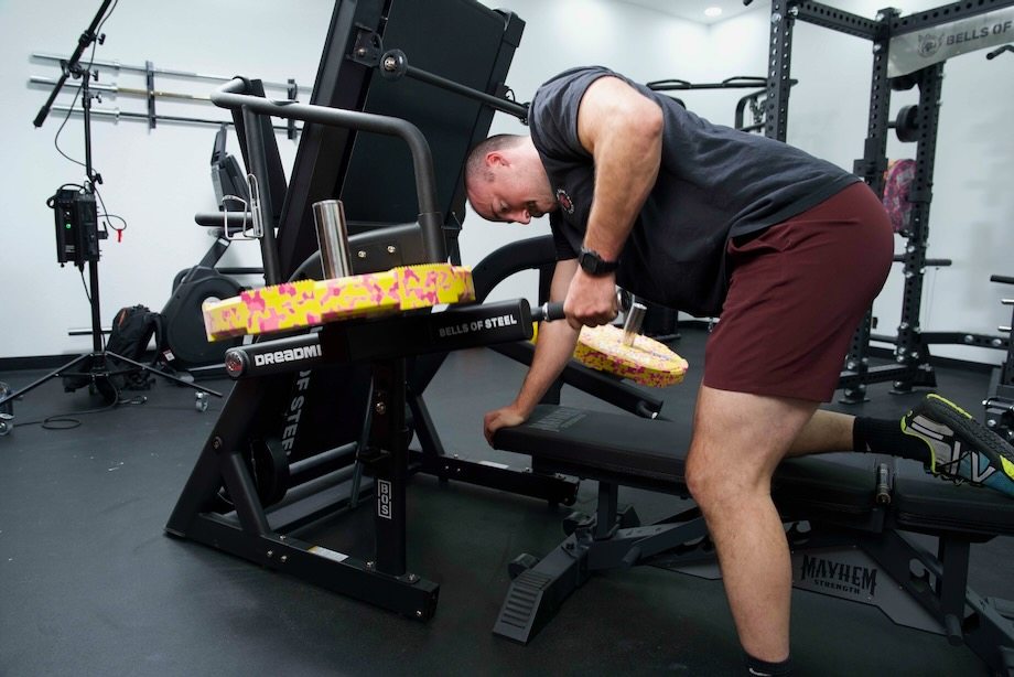 Coop lifts a huge weight plate with one arm while leaning on the bench of a Bells of Steel Dreadmill.