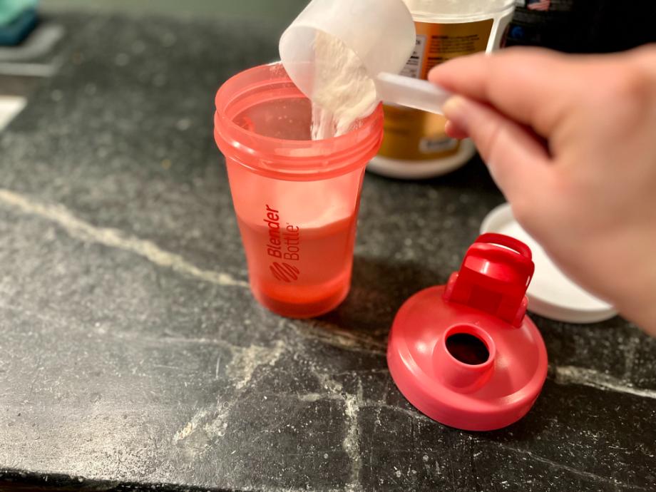 A person's hand pours a scoop of protein powder into the Blender Bottle Classic V2