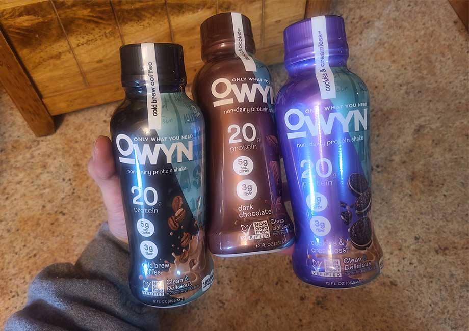 A hand holding three different flavors of OWYN protein shake. The bottles are black, brown, and purple.