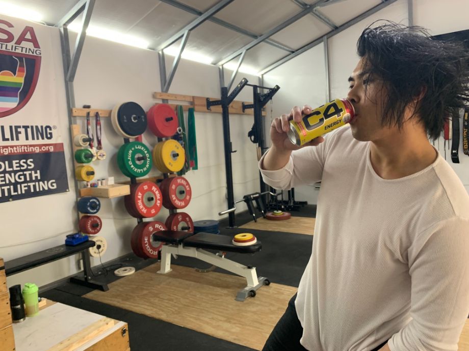 Person wearing a white long-sleeved shirt drinking a can of C4 Energy drink in a gym 