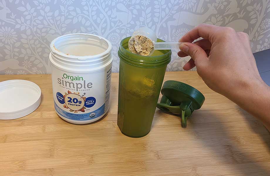 A person pours a scoop of Orgain Simple Plant Protein into a shaker cup.