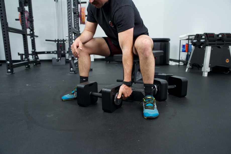 Coop adjusts an Eisenlink Adjustable Dumbbell while sitting on a weight bench.