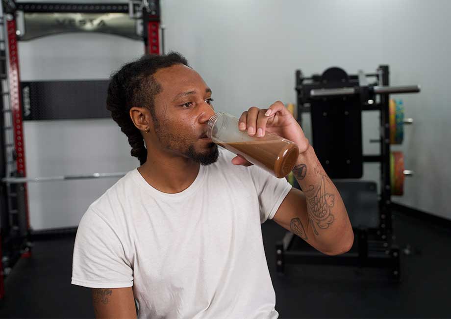 A man with braids and a beard drinking a brown protein shake in a gym setting