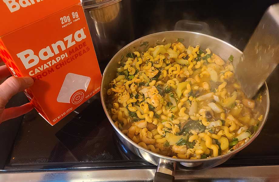 A Banza box is held next to a pot of cooking pasta.