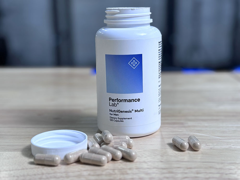 Close-up of open white and blue bottle of Performance Lab NutriGensesis Multi for Men, sitting behind about a dozen loose capsules sitting on a table next to the bottle's lid.