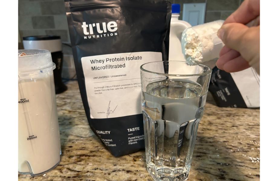 A person dumping a scoop of True Nutrition whey protein isolate into a glass. The bag of protein powder is sitting on the counter next to the glass.