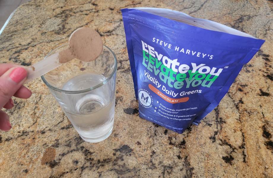 A scoop of L’Evate You Greens Powder being poured into a glass of water.