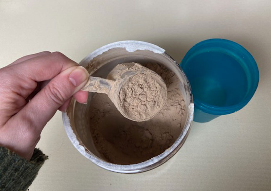 A hand lifting a scoop of Purely Inspired Organic Protein powder from the container.