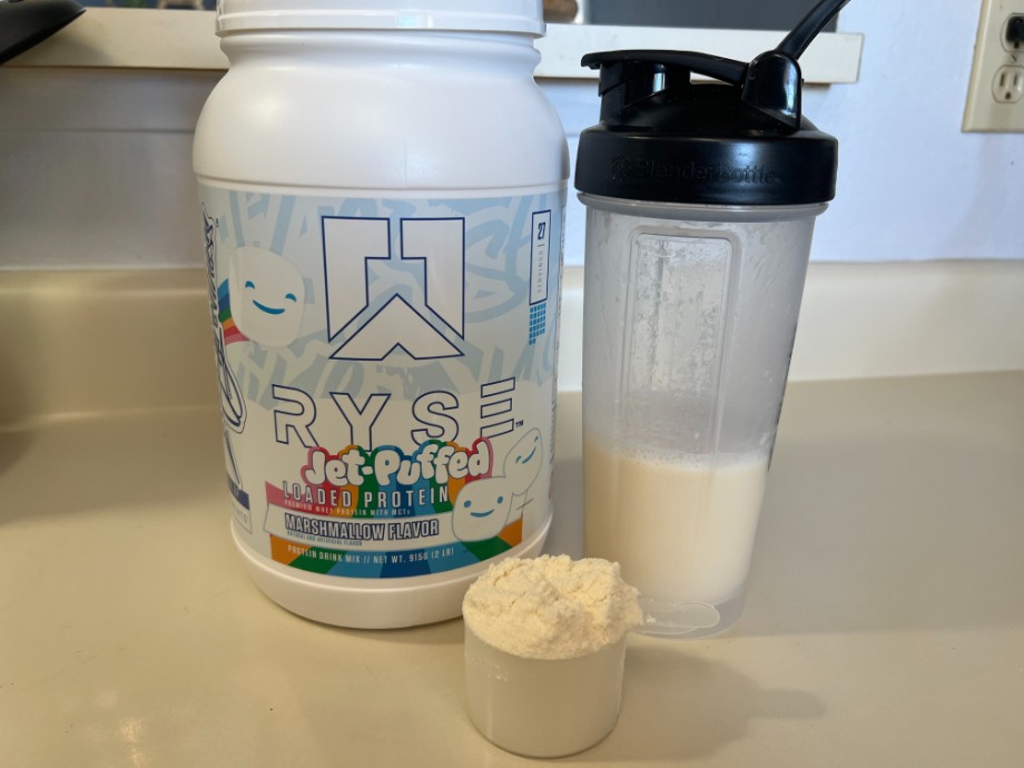 A scoop of RYSE Protein Powder is next to the container and a shake.
