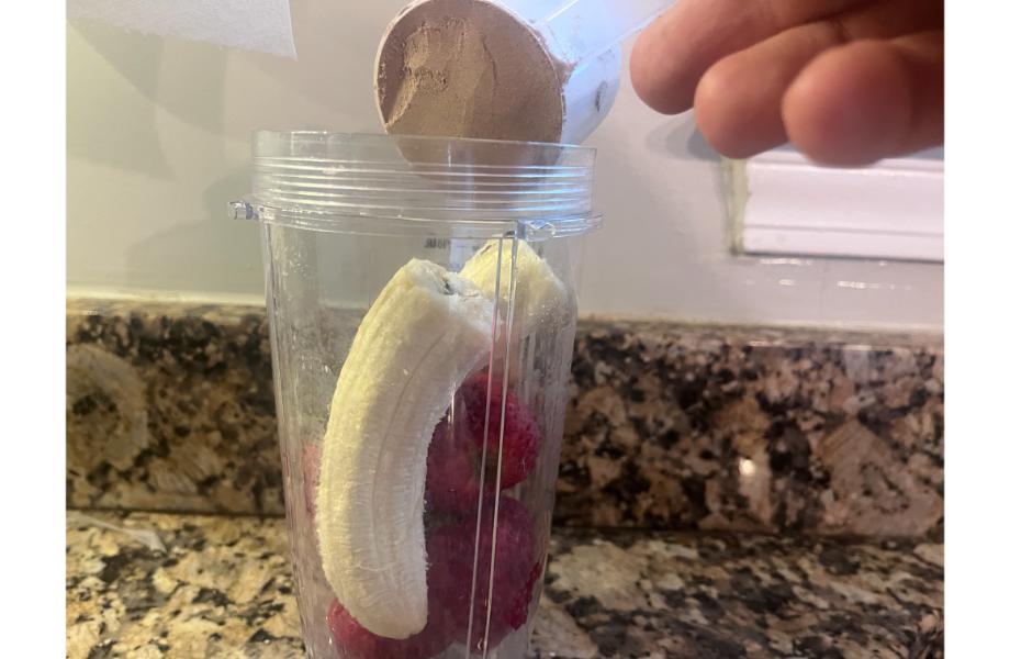 Someone pouring a scoop of Sports Research protein powder into a blender with fruit
