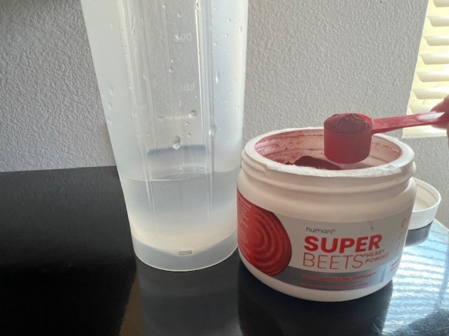 A person holds up a scoop of SuperBeets before mixing it into a glass of water.