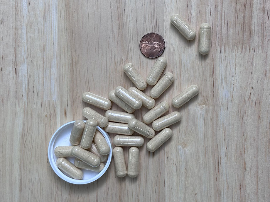 Closeup of over 20 Transparent Labs Multivitamin capsules, filled with a cream-colored powder and sitting amongst the product bottle's lid and a copper penny as a size comparison.