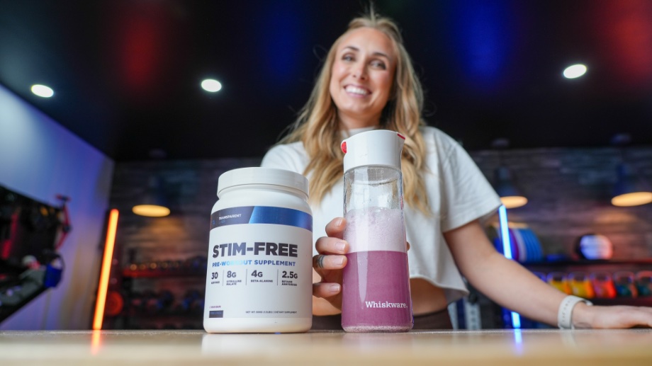 A woman can be seen smiling behind a shake and container of Transparent Labs Stim-Free Pre-Workout.
