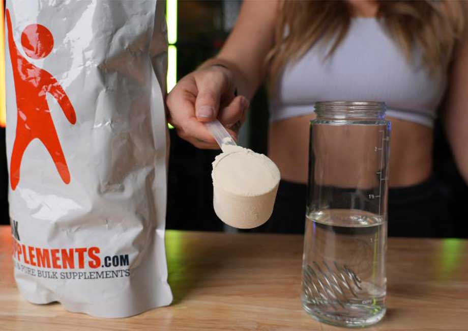 A woman holds up a scoop of Bulk Supplements Soy Protein Isolate next to a shaker glass of water.
